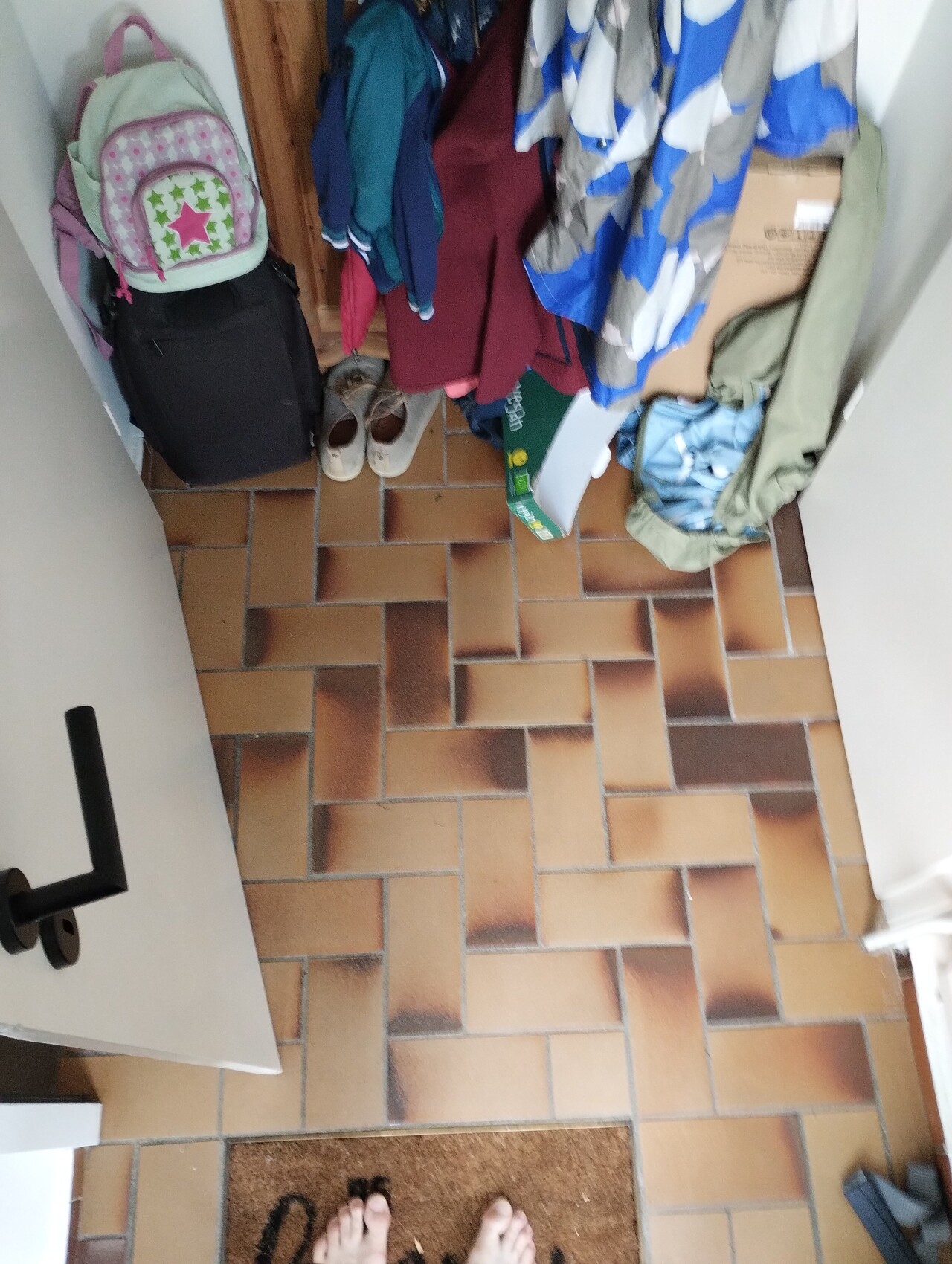 Flur-Eingang: Jacken am Haken, Schuhe, Rucksack, Karton, braune Fliesen, barfuß am Boden