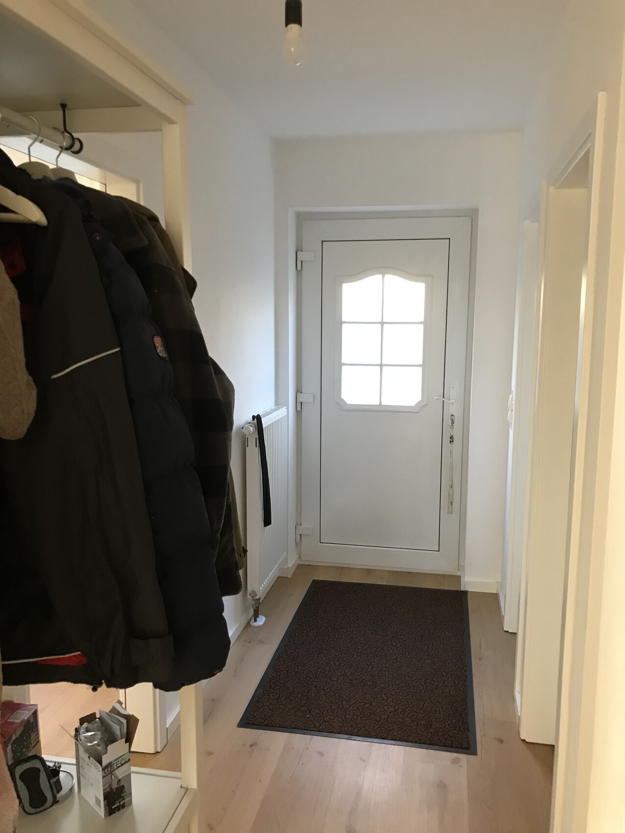 Flur-Eingang: links Garderobe mit Mänteln, mittig weiße Tür mit Fenster, braune Fußmatte.