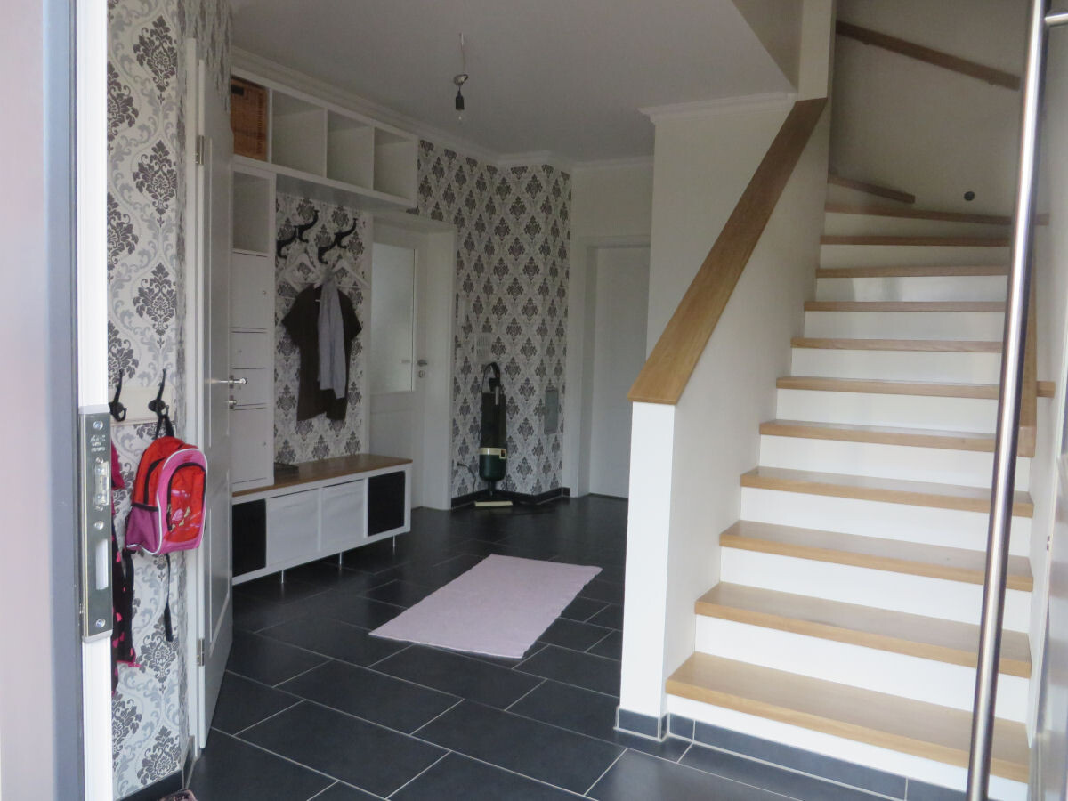 Eingangsbereich mit Treppe rechts, Garderobe, gemusterter Tapetenwand und pinkem Teppich.