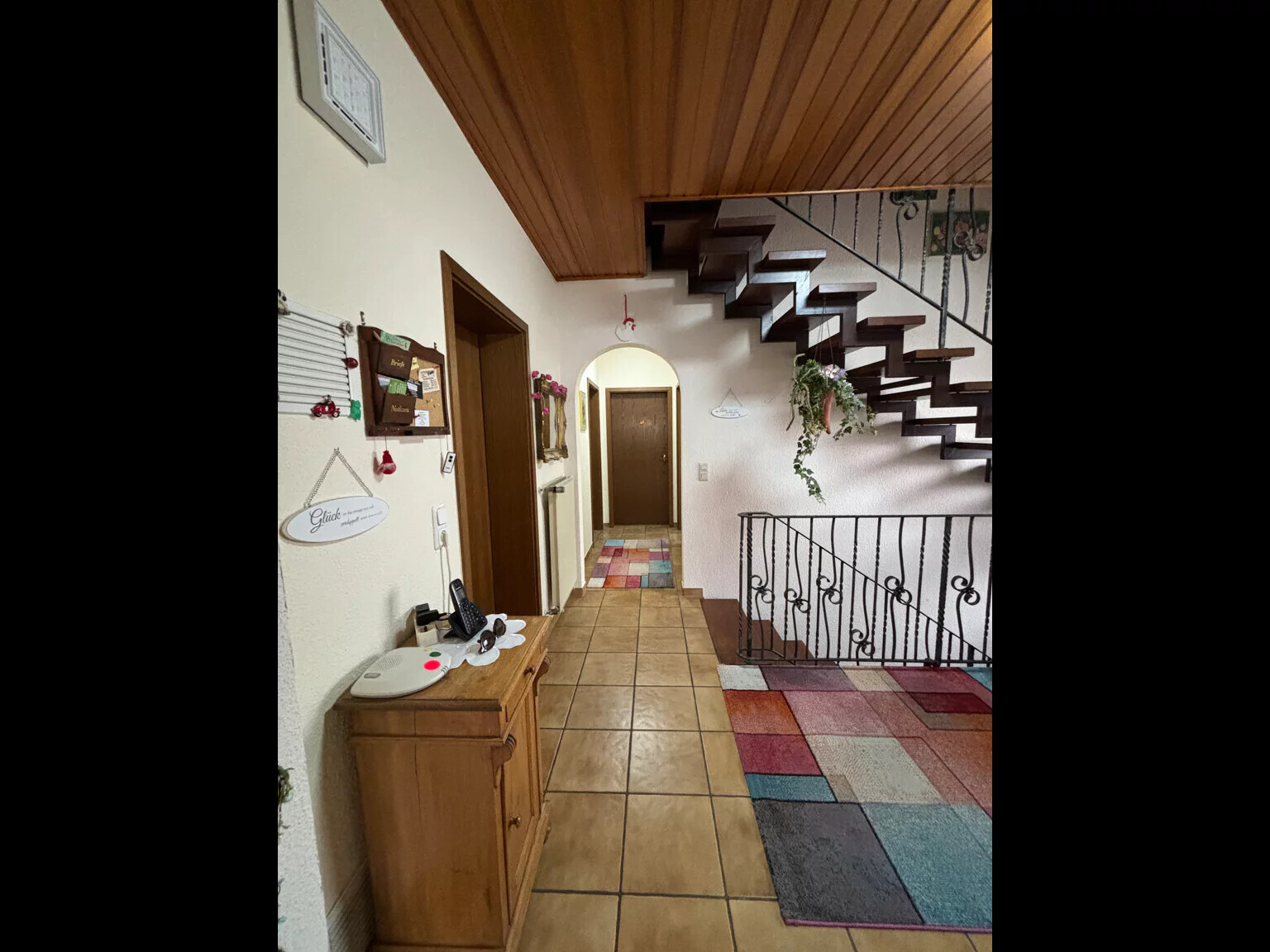 Flur mit Holzboden und Holzdecke, Treppe, schmiedeeisernes Geländer, bunte Teppichfliesen, Wanddeko.
