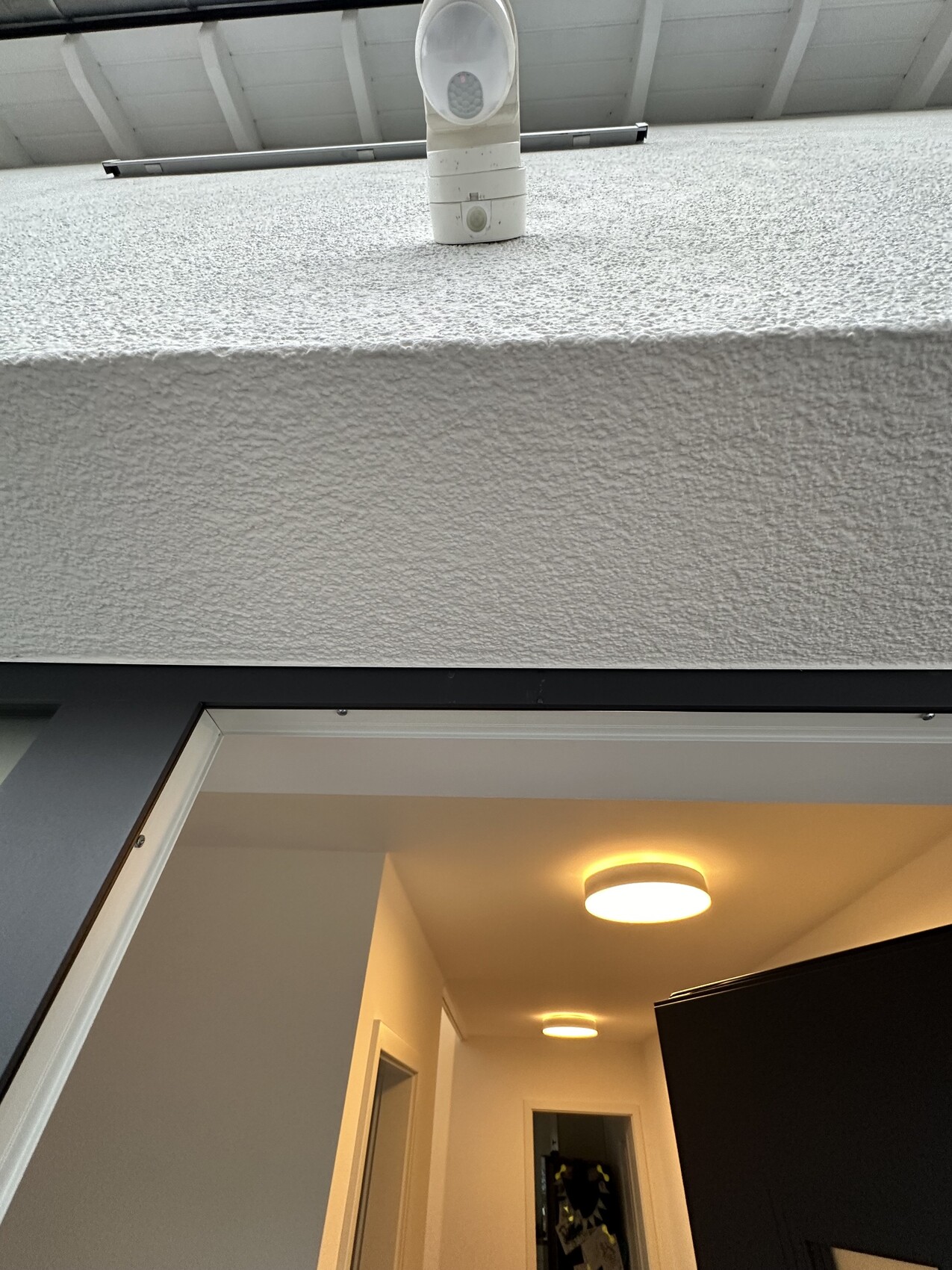 Weiße Außenleuchte mit Bewegungsmelder an rauer Wand; Tür zum hellen Innenflur.