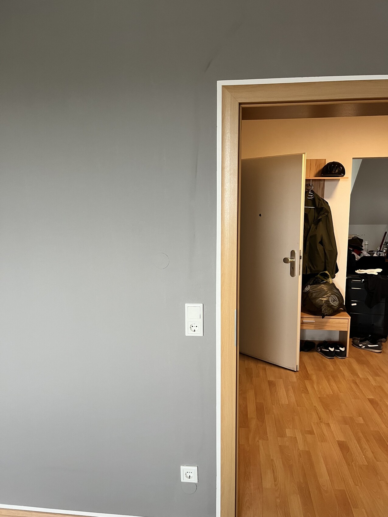 Offene Zimmertür in grauer Wand führt zu Flur mit Garderobe, Schuhen und Helm auf Bank.