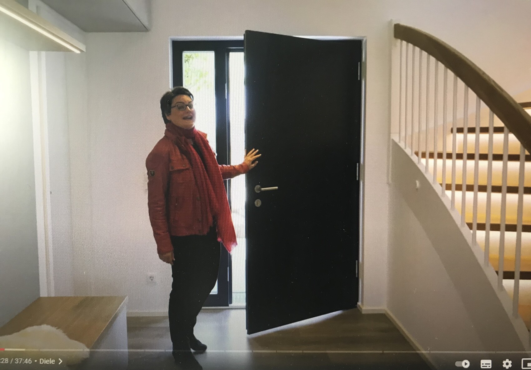 Eine Person in roter Jacke und Schal steht im Flur vor offener schwarzer Tür; Treppe rechts.