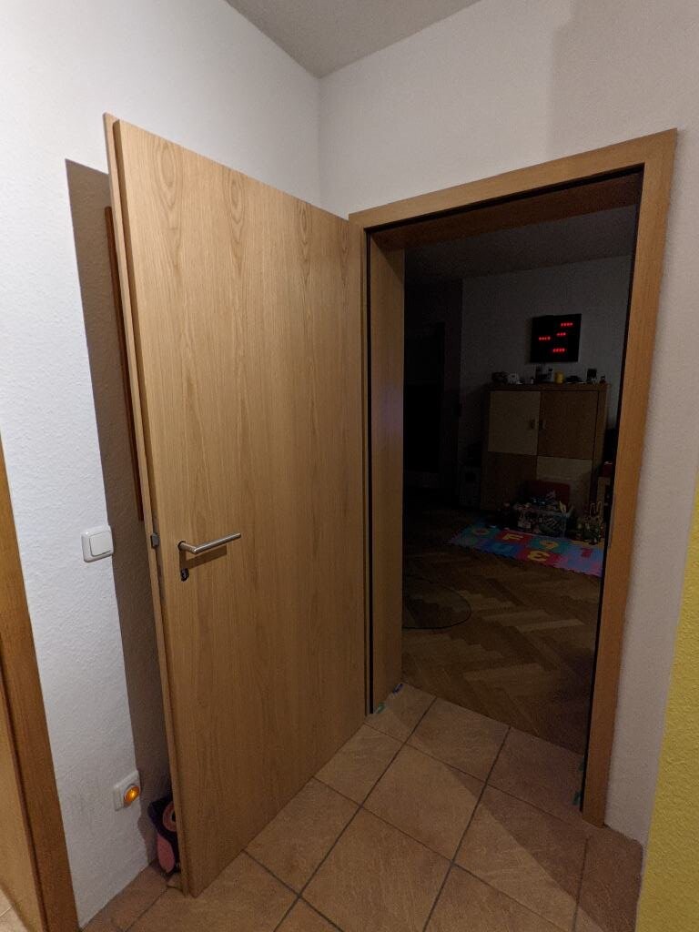 Offene Holztür im Flur führt in Wohnraum mit Spielbereich im Hintergrund.