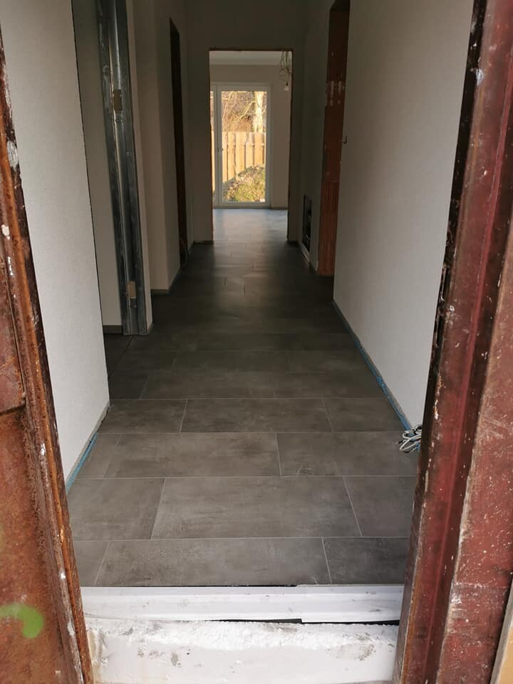 Langer Flur in Renovierung mit grauen Bodenfliesen, weißen Wänden und Licht am Ende.