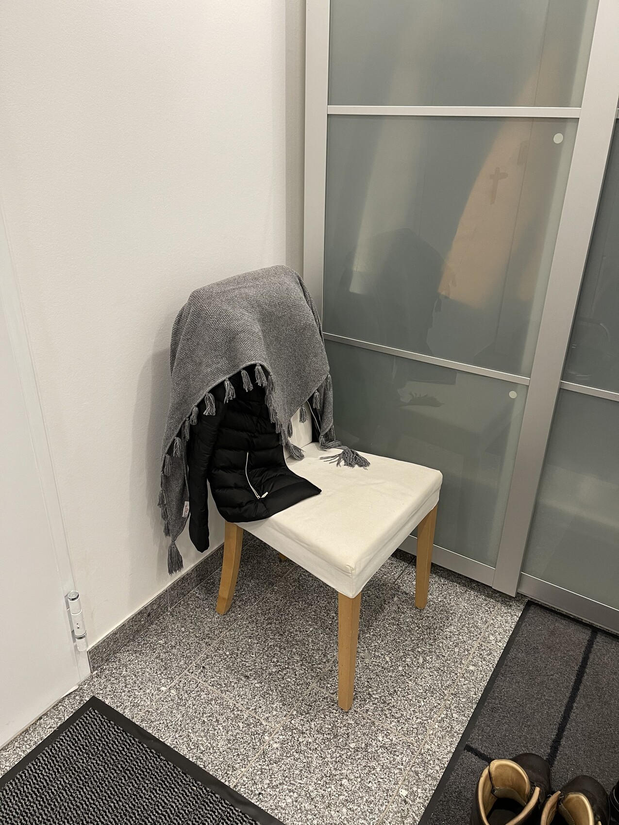 Stuhl mit weißem Sitzkissen, graue Fransen-Decke über schwarzer Jacke; Glas-Schrank dahinter.