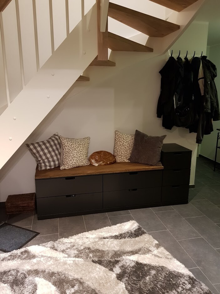 Unter der Treppe: Sitzbank mit Kissen, schlafende Katze, Garderobe mit Jacken, schwarze Kommode.