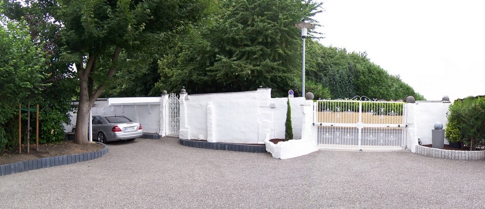 Weiße Mauer mit Tor und Einfahrt; links geparktes silbernes Auto, Garten dahinter.