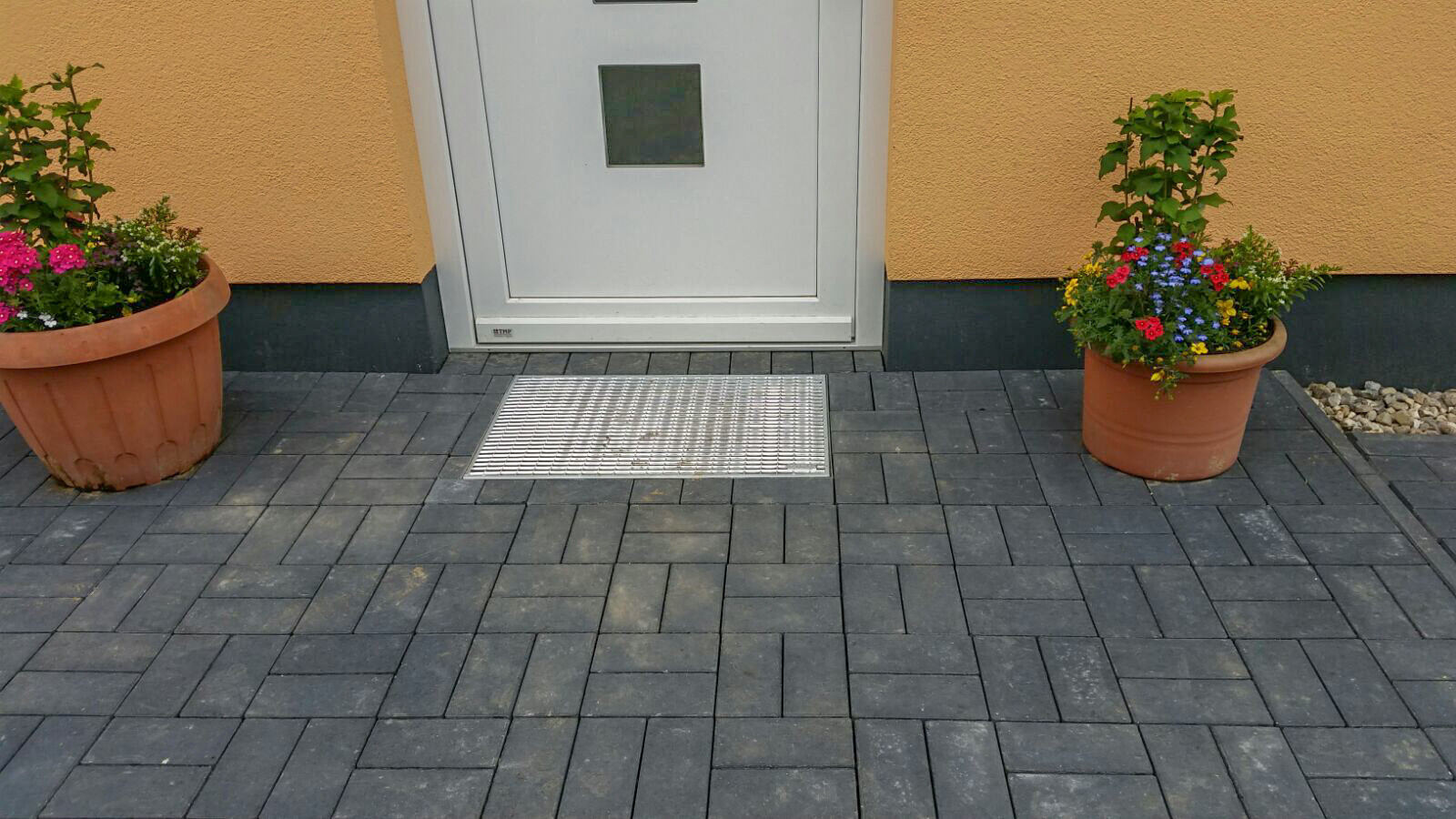 Eingangsvorbereich mit grauen Pflastersteinen, weißer Tür, Metallgitter und zwei Blumenkübeln.