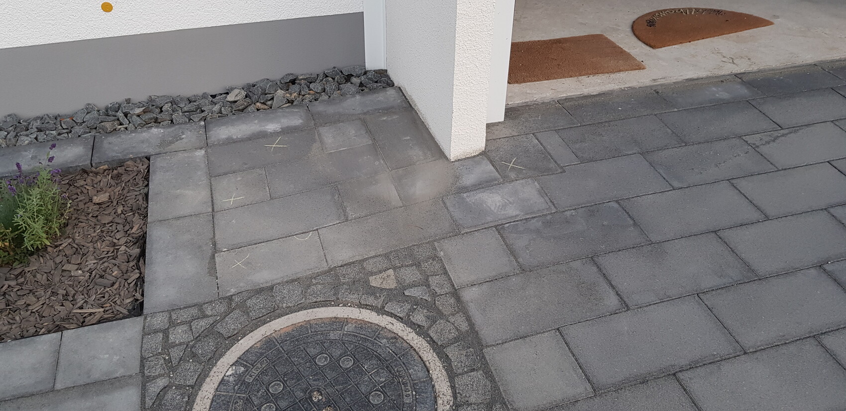 Grauer Pflasterweg vor dem Haus, Lavendelbeet links, runder Kanaldeckel, Kreidemarkierungen auf Pflastersteinen.