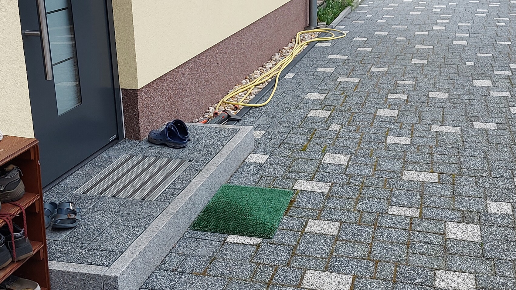 Eingangsbereich: graue Tür, Schuhregal, Schuhe, gelber Schlauch und gepflasterter Weg.
