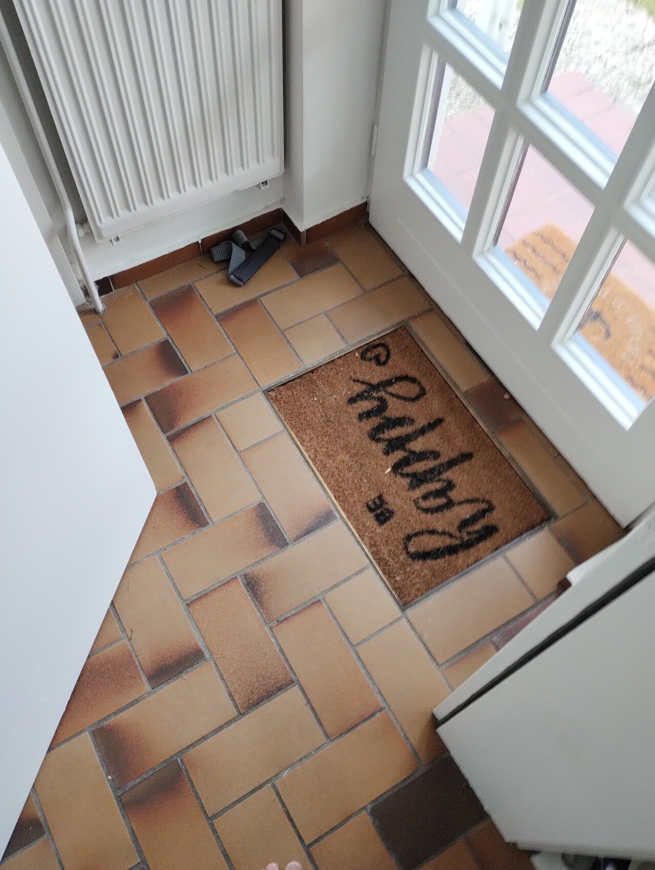 Flur mit terrakottafarbigen Fliesen, weiße Tür mit Glasfenstern, Willkommensmatte 'Hello'.