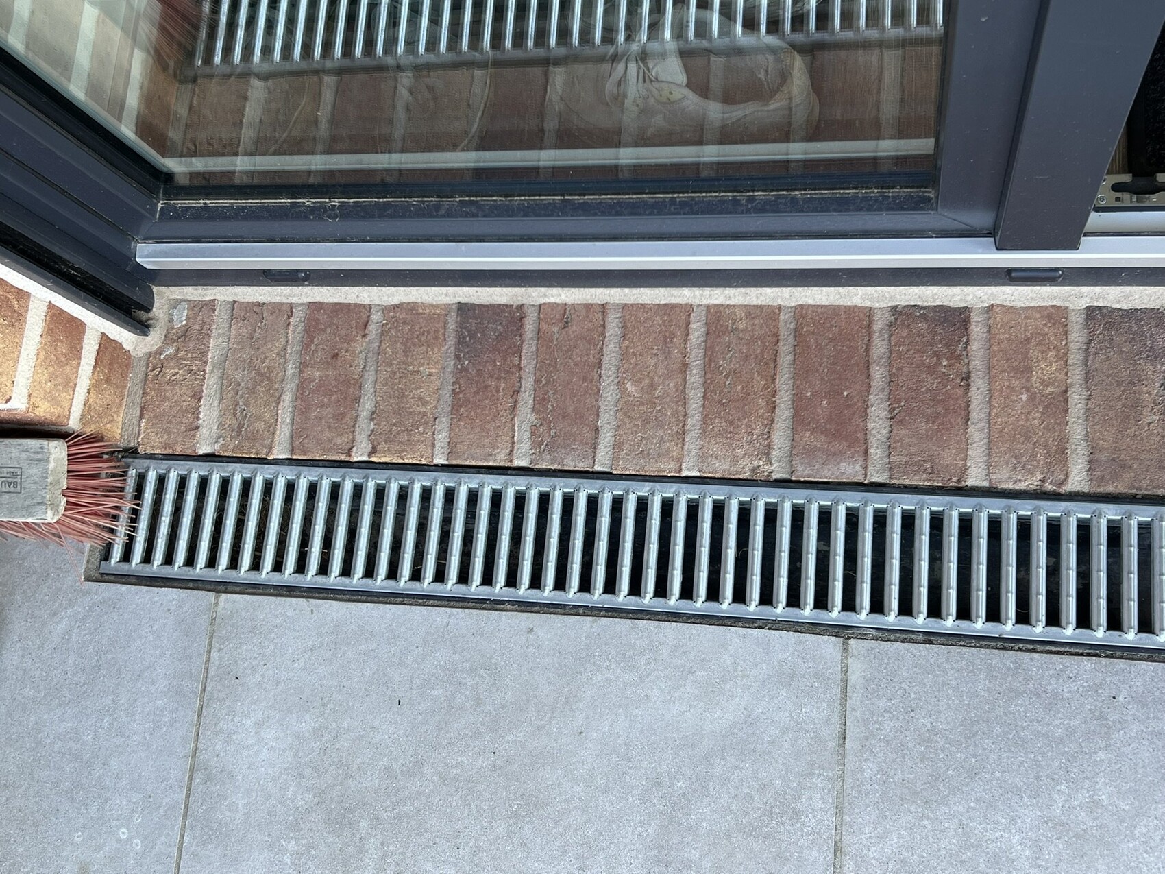 Außenbereich vor einer Glastür; roter Ziegelboden, lange metallene Abflussrinne, linke Bürste.