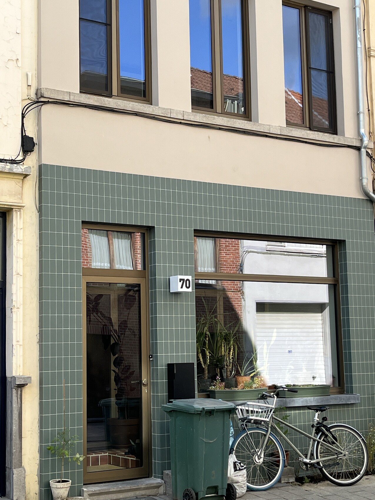 Gebäudevorderseite mit grüner Fliesenverkleidung, Tür Nr. 70, Fahrrad und Mülltonne davor.