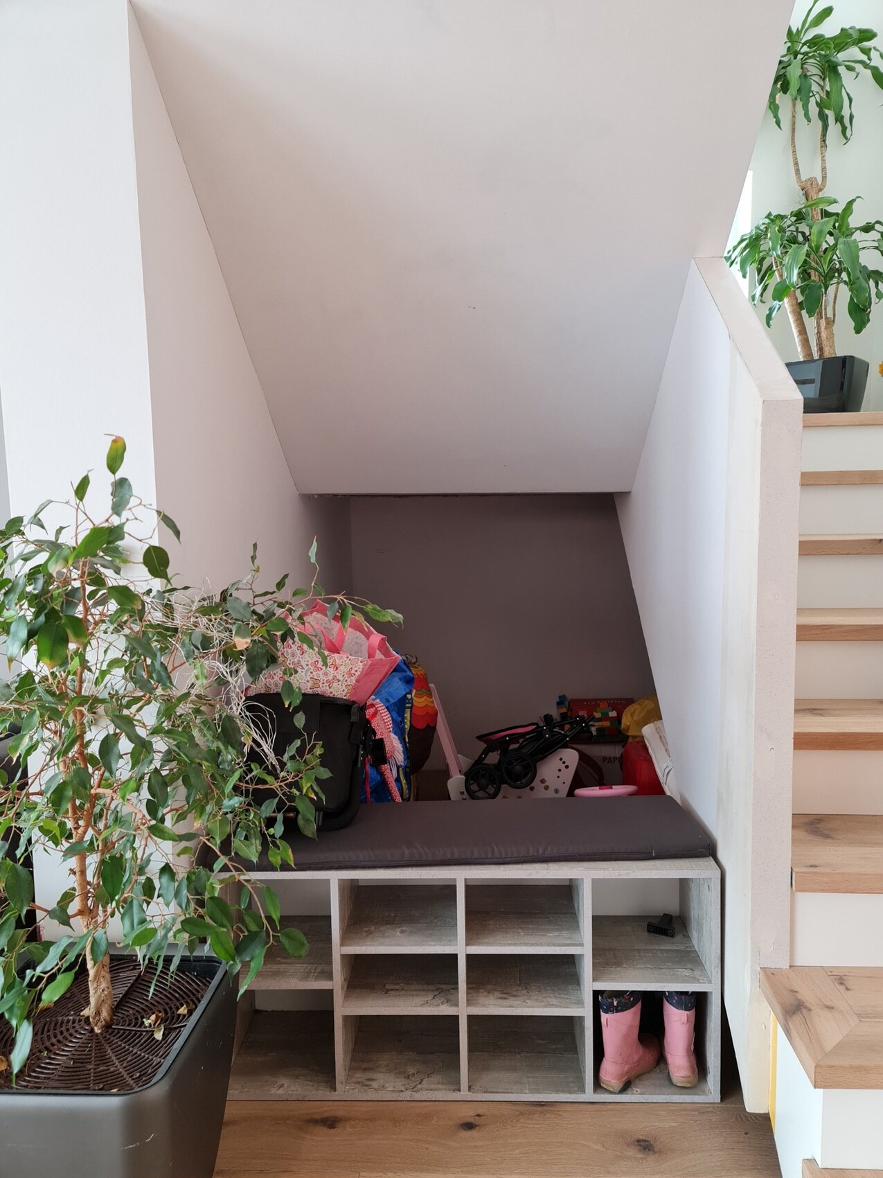 Sitzbank unter der Treppe mit Fächern, Schuhen, Spielzeug; links eine große Pflanze.