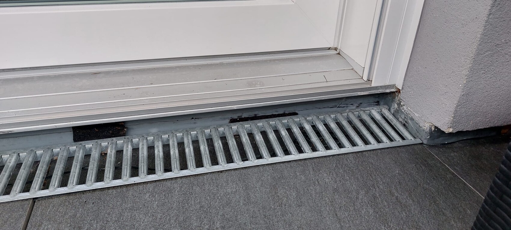Schiebetür mit darunterliegendem langen Metallabflussgitter vor grauer Außenwand.