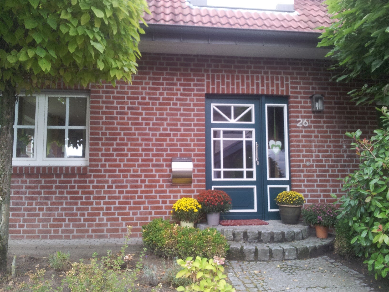 Frontansicht eines roten Backsteinhauses mit blauer Tür, Fenster links und bunten Topfpflanzen.