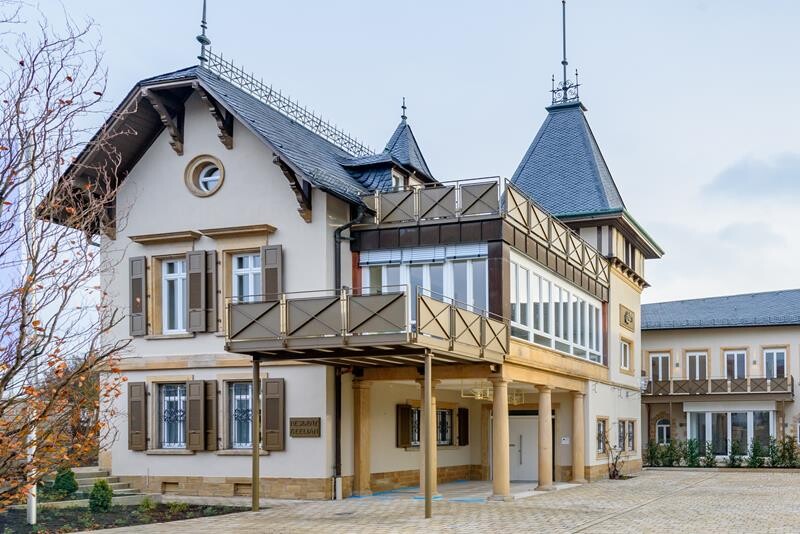 Zweistöckige Villa mit Holzfensterläden, umlaufendem Balkon, Turmspitze und verglaster Veranda.