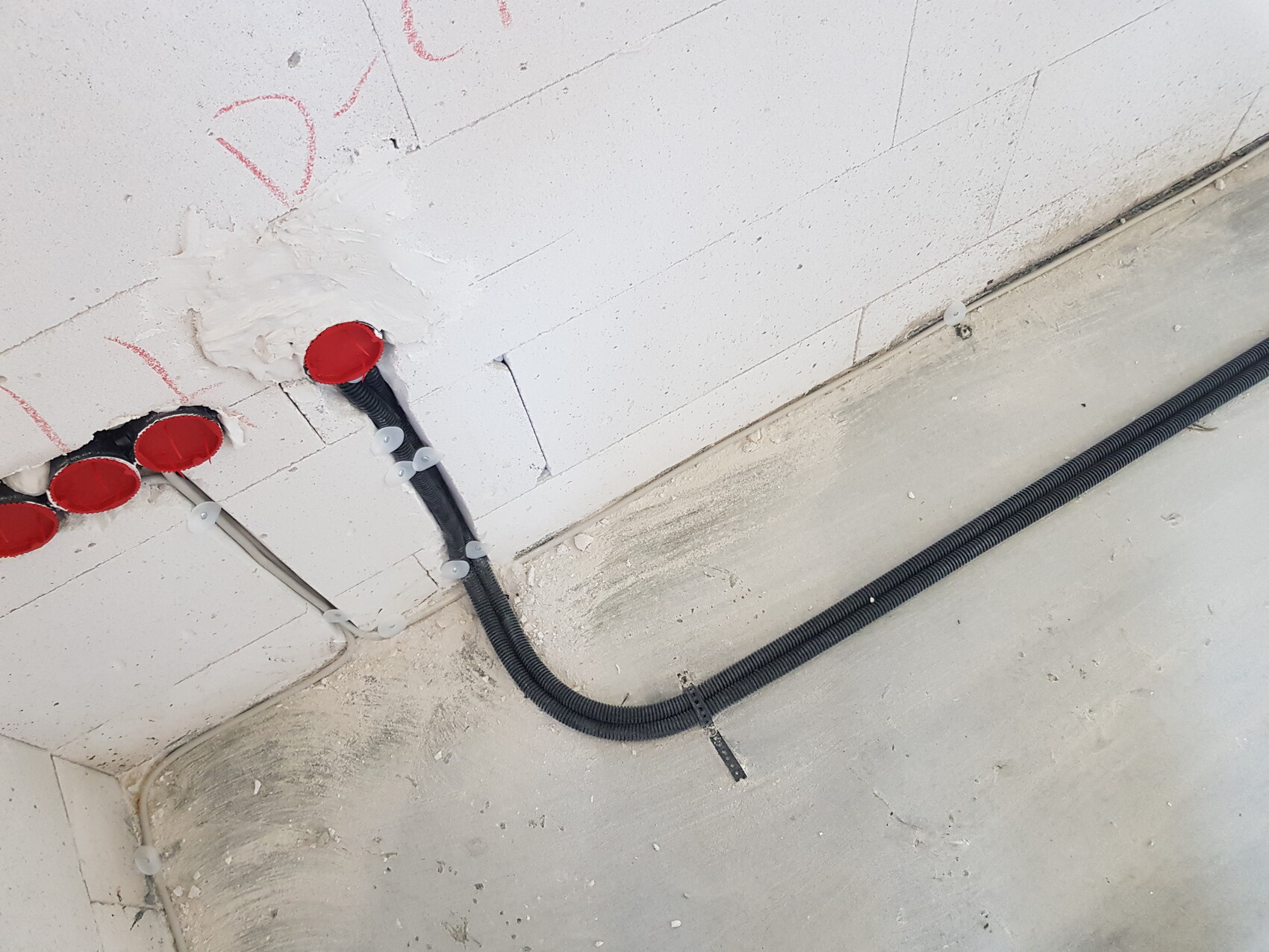 Bauwand mit roten Abdeckkappen in Rohrdurchführungen und schwarzem Kabelrohr am Boden.