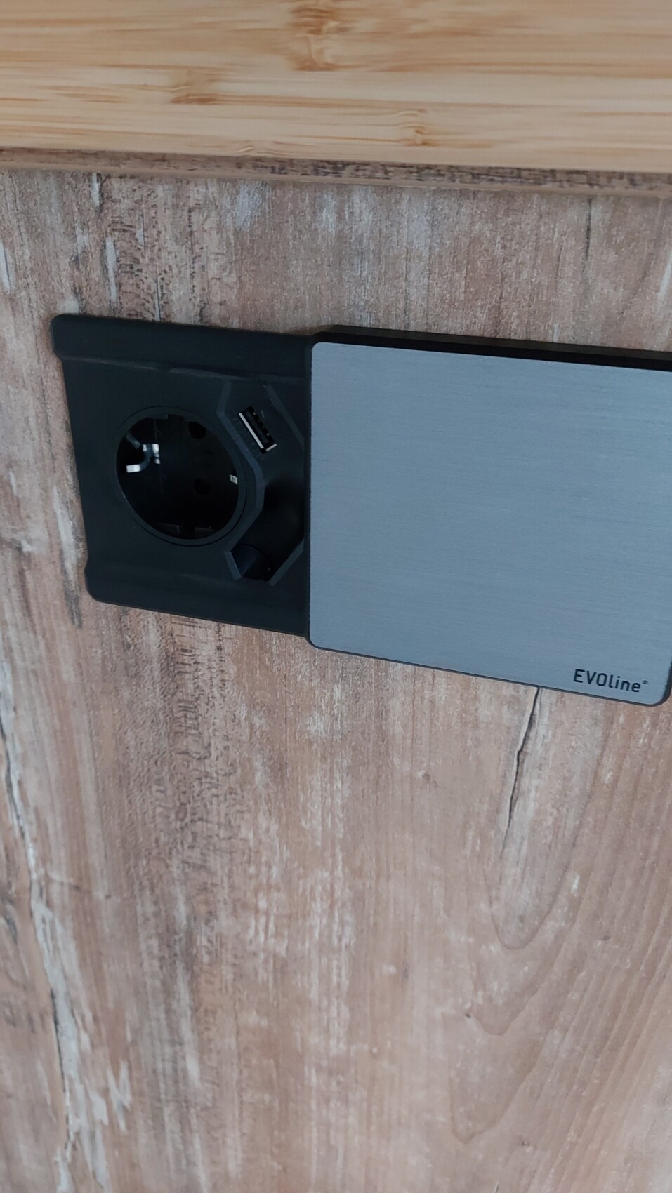 Schwarze Wandsteckdose mit USB-Anschluss neben grauem EVOLine-Panel an Holzwand.