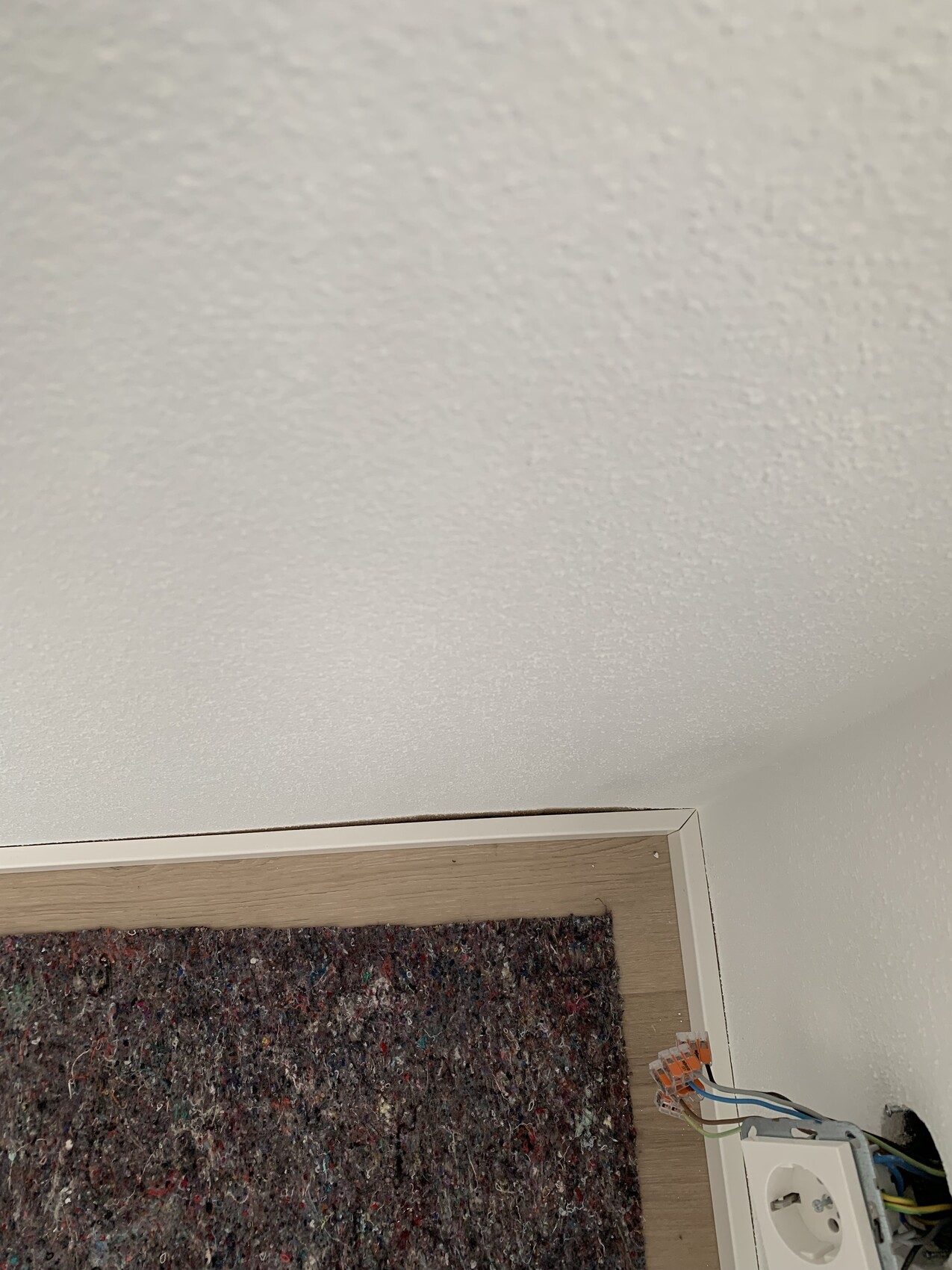 Eckansicht: bunter Teppich, Holz-Sockel, weiße Wand, freiliegender Stromanschluss mit Kabeln.