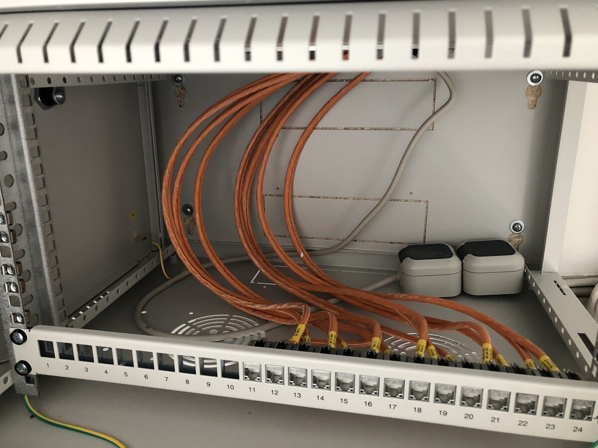 Netzwerk-Schrank mit orangefarbenen Kabeln am Patchpanel und zwei weißen UPS-Kästen.