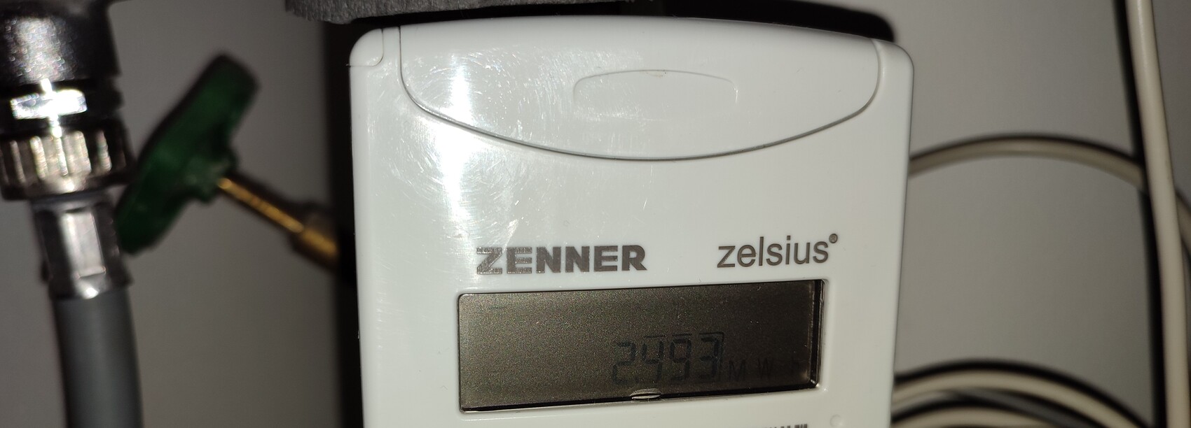 Weißes ZENNER zelsius Messgerät mit Display, links Rohrleitungen und Ventil sichtbar.