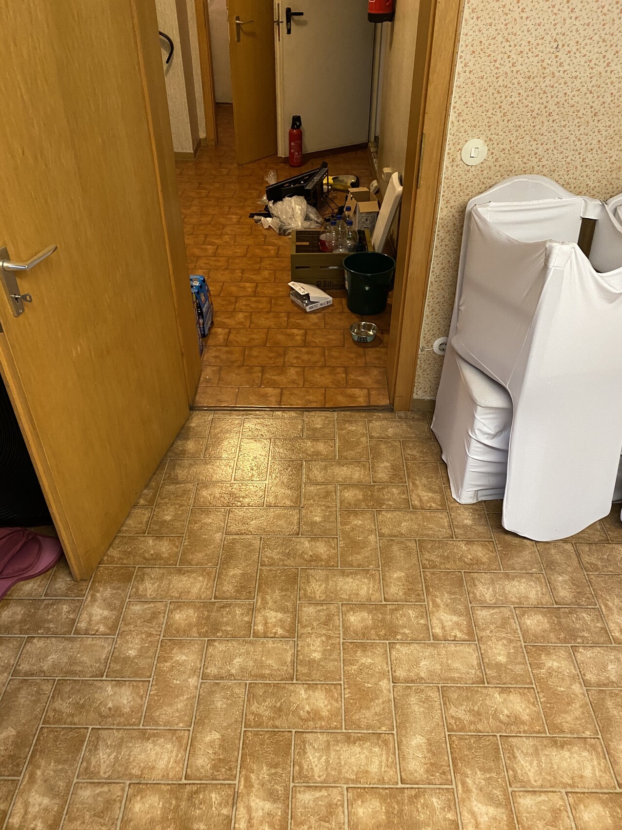 Offener Flur mit Unordnung: Kartons, Flaschen, Eimer, Hunde-Napf; rechts weißes Sofa.