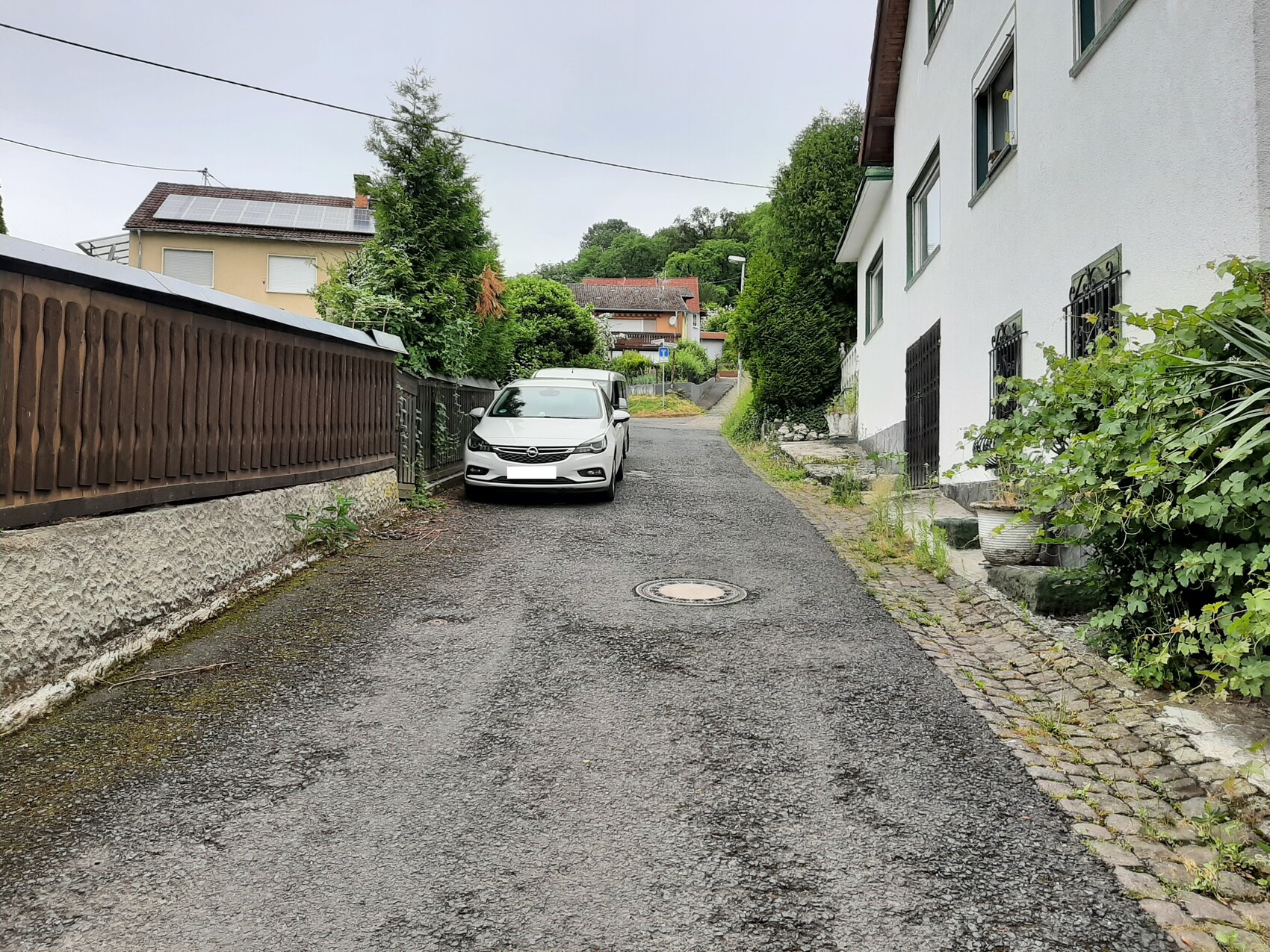 Schmale Wohnstraße: links geparktes Auto, Holzzaun, rechts weißes Haus mit Fenstern und Pflanzen.