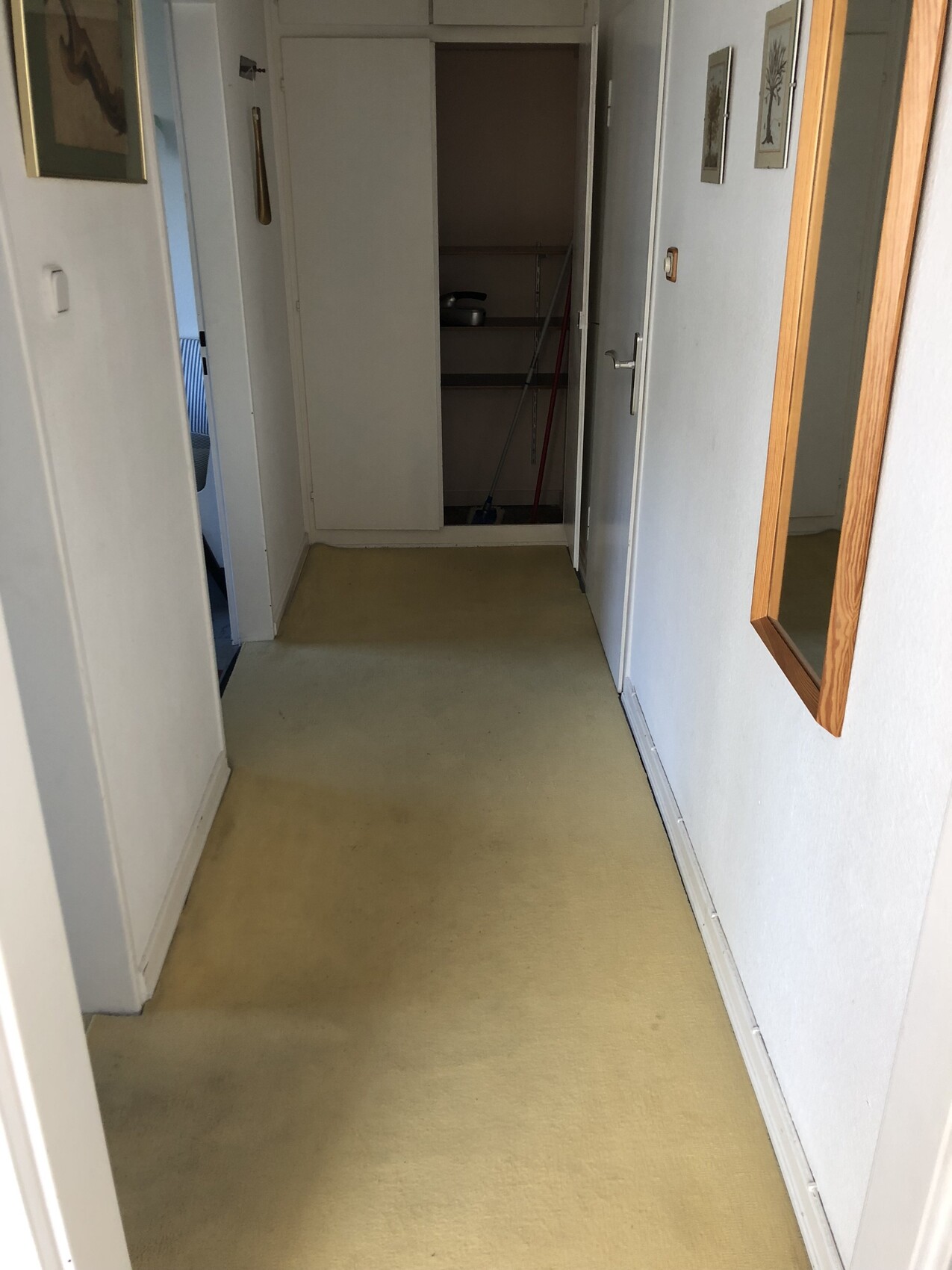 Heller Flur mit gelbem Boden; am Ende geöffnete Abstellkammer mit Besen/Mopp; Spiegel rechts.