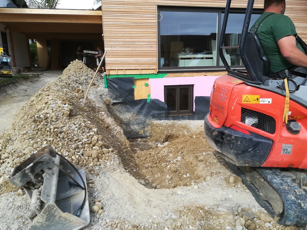 Minibagger neben Baugrube vor Holzhaus, Erdhaufen, Bauarbeiten im Hintergrund.