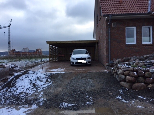 Weißes Auto steht unter Carport neben Haus; Schnee und Matsch auf der Zufahrt, Baukran im Hintergrund.