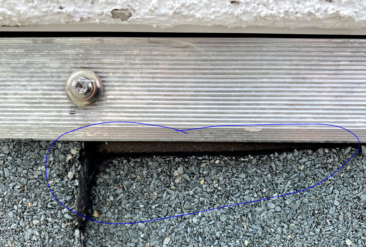 Aluminium-Haustürschwelle mit Schraubenkopf; darunter Kiesboden sichtbar.