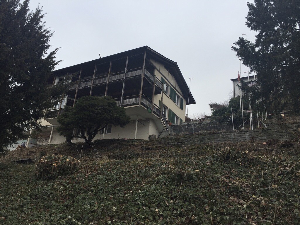 Zweistöckiges Haus mit Holzveranda auf Hanglage, umgeben von Bäumen und Steinmauern.