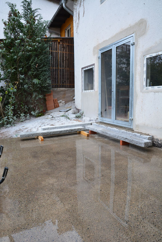 Außenansicht eines Hauses mit Glas-Schiebetür; Brücke aus Beton auf Holzstützen über nasser Fläche.
