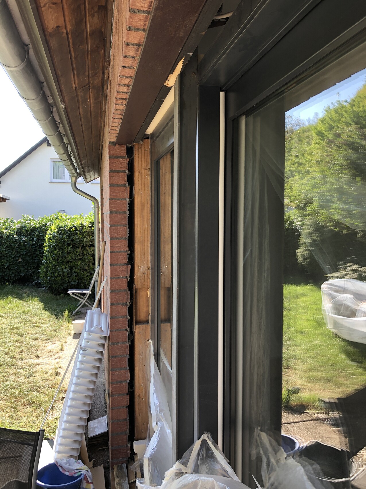 Außenansicht: Ziegelwand mit freigelegtem Holzrahmen, große Glasfront, Bauutensilien im Garten.