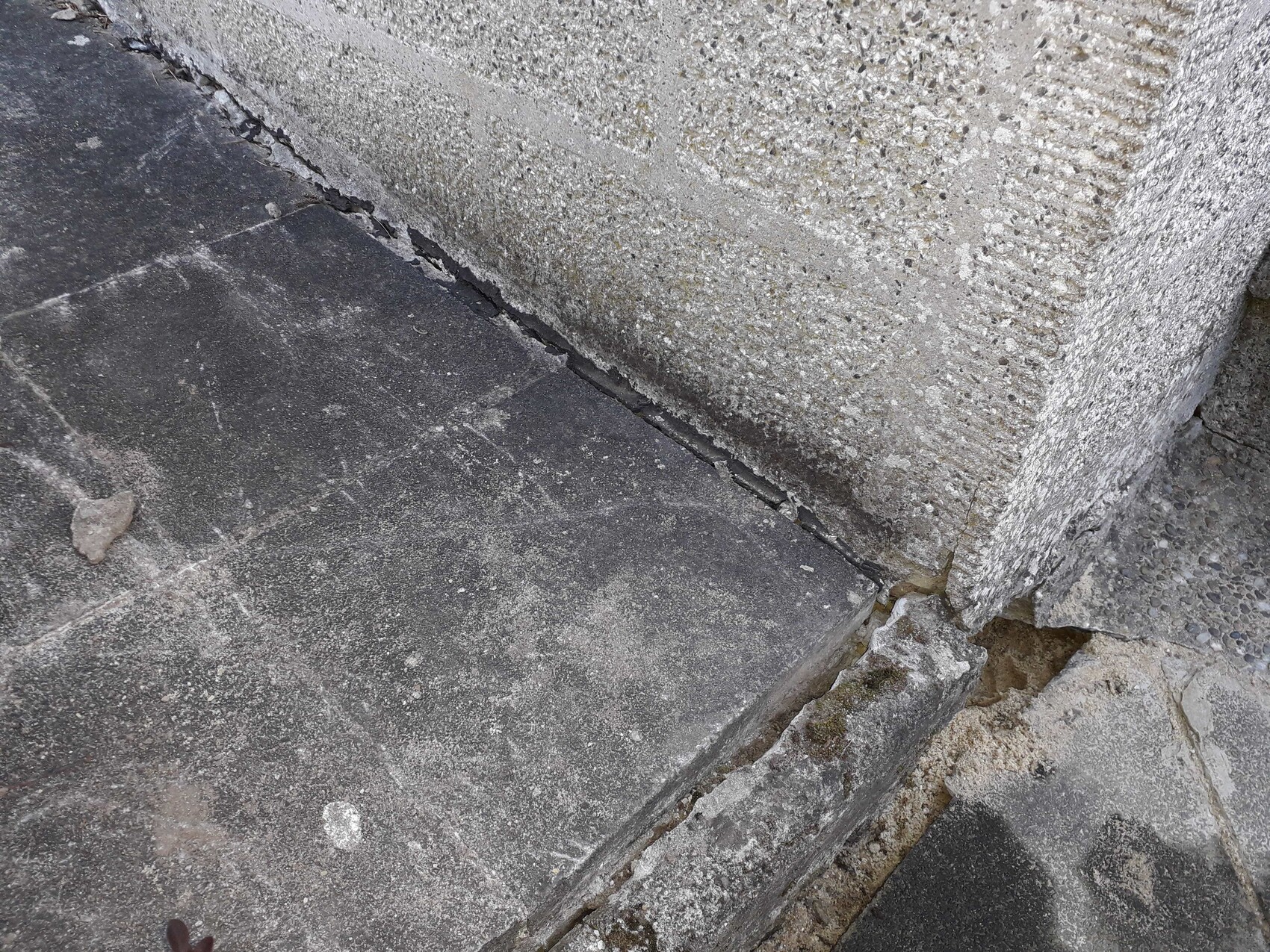 Nahaufnahme grauer Steinplatten mit Rissen, neben rauer Steinmauer.