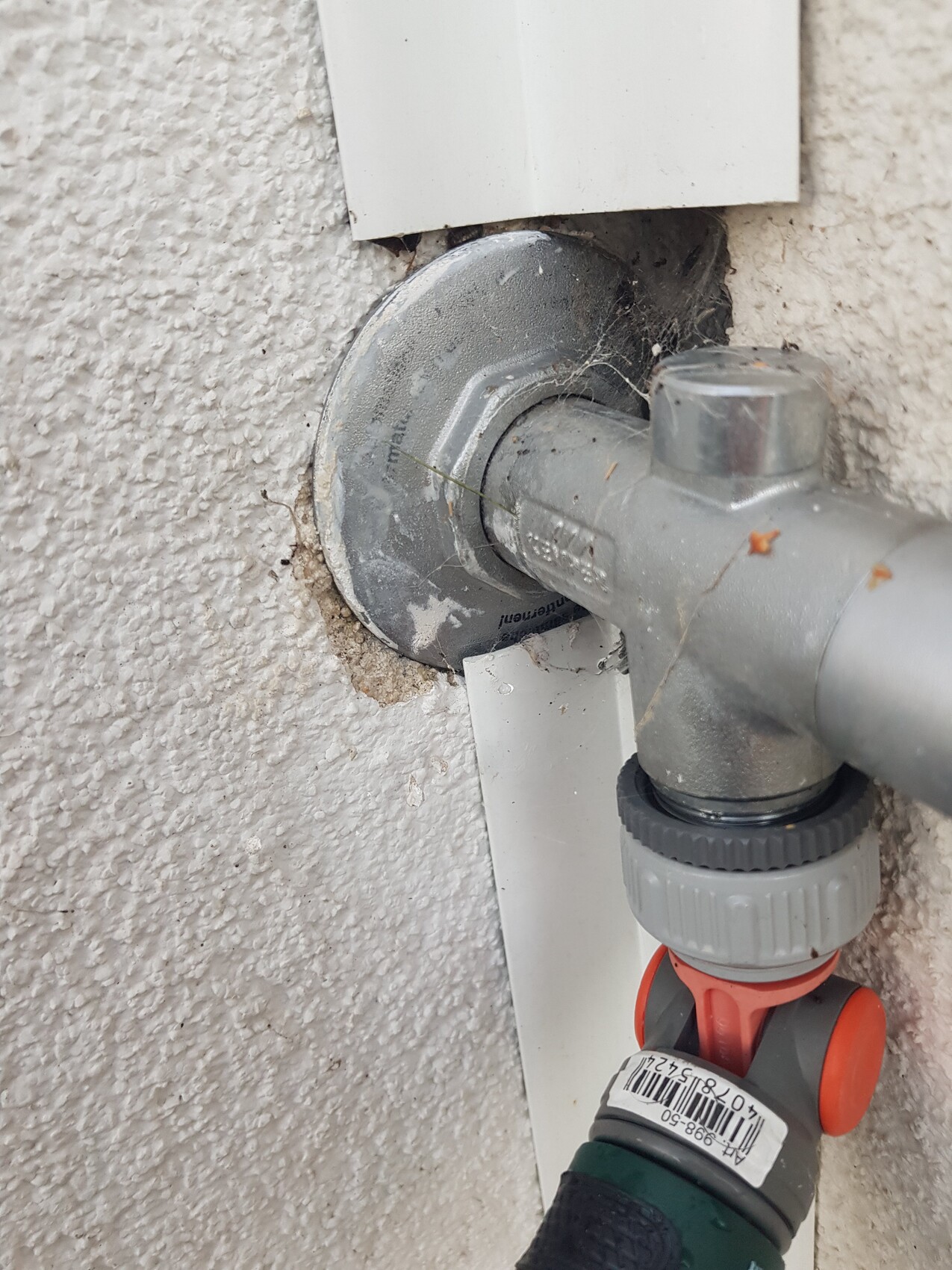 Außenwasseranschluss am Mauerwerk mit Metallventil, grauem Griff und orangefarbenem Knopf.