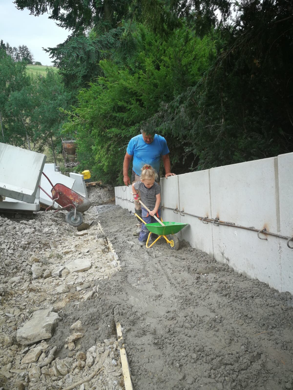 Ein Mann in blauem Shirt hilft einem Kind beim Graben mit grünem Kippwagen neben einer Betonwand.