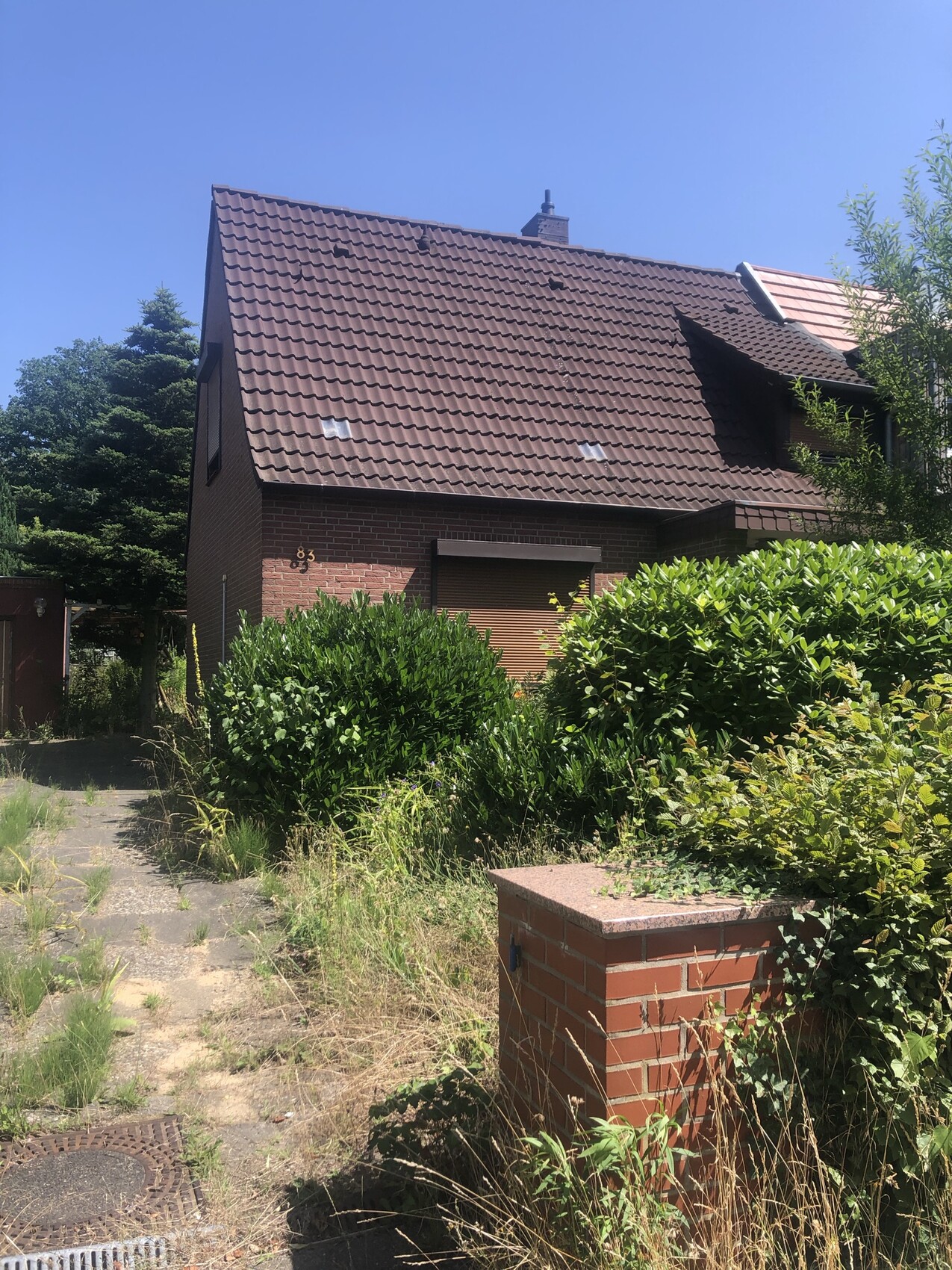 Zweistöckiges rotes Ziegelhaus mit braunem Satteldach, umgeben von dichten Sträuchern.