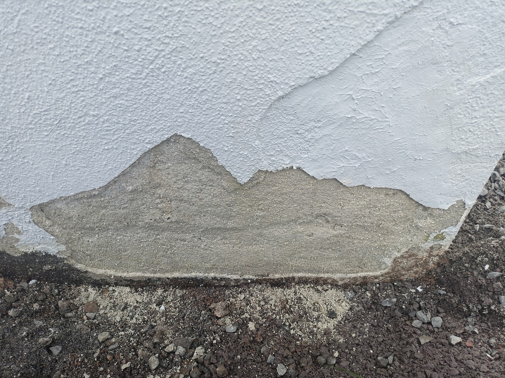 Wand mit abblätterndem Putz; darunter grauer Beton, Boden aus Kies und Geröll.