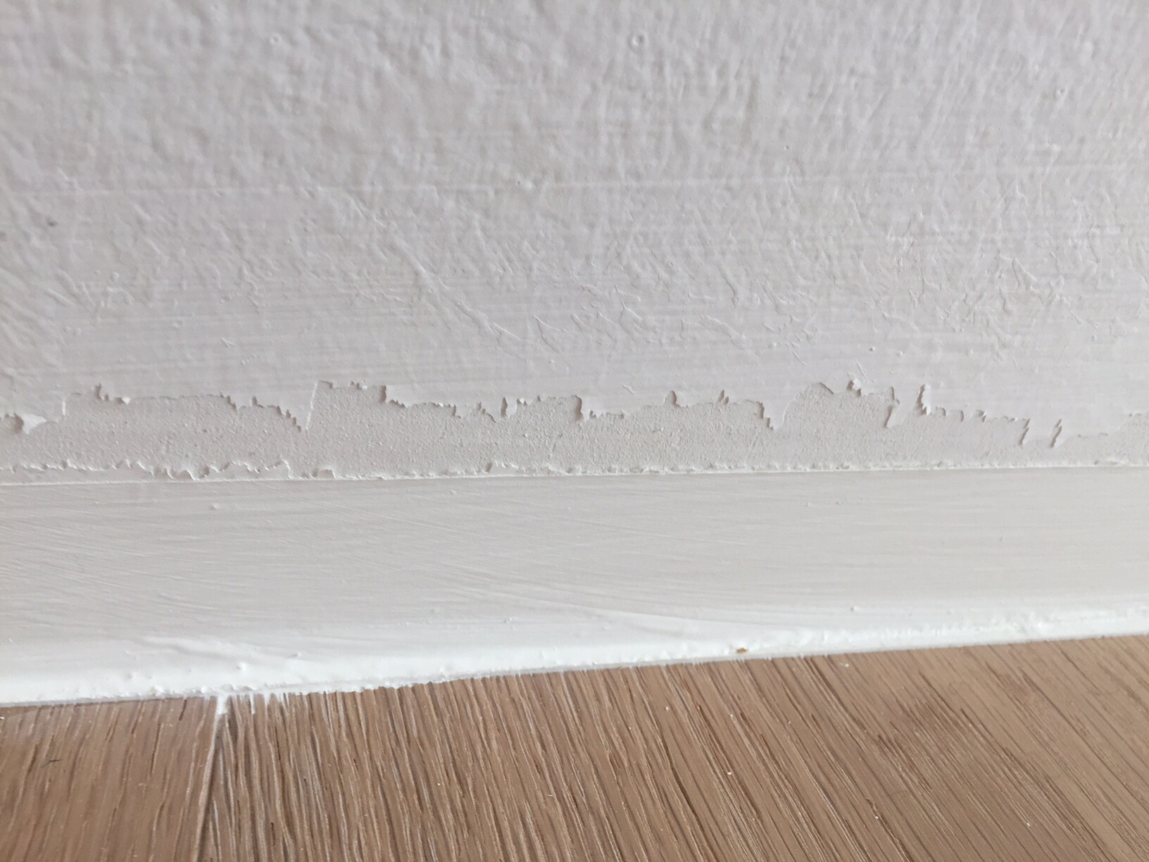 Weiße Wand mit abblätternder Putzschicht über Sockel, Holzboden darunter