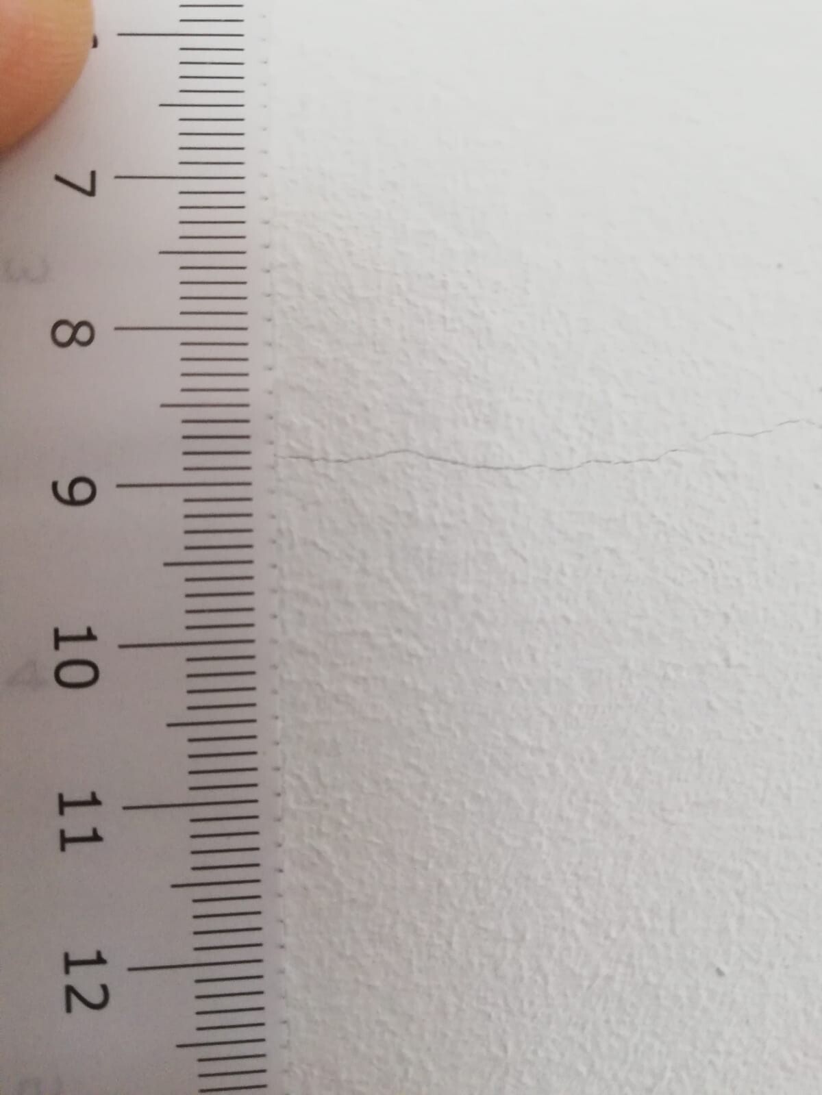 Nahaufnahme eines Lineals an einer weißen Wand; Zentimeter-Skala 6-12 cm, Finger sichtbar