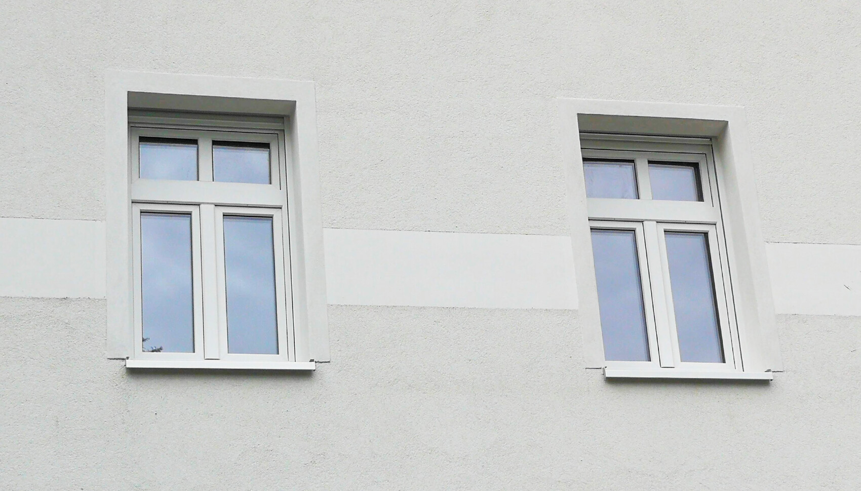 Zwei weiße Fenster mit Rahmen in einer hellgrauen Putzfassade; spiegeln blauen Himmel.
