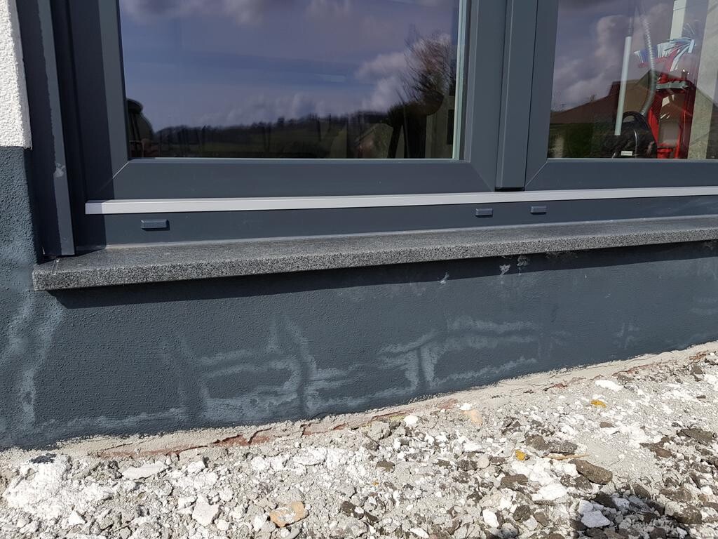 Außenansicht: graue Fensterrahmen mit Fensterbank, dunkle Wand und Kies davor.
