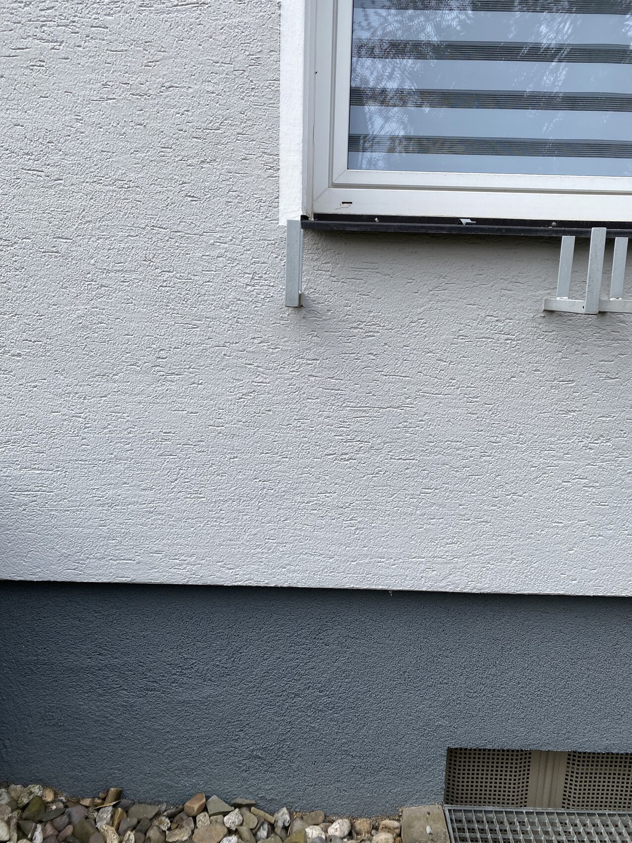 Hellgraue Außenwand mit Fenster, dunklem Sockel und Kiesboden.