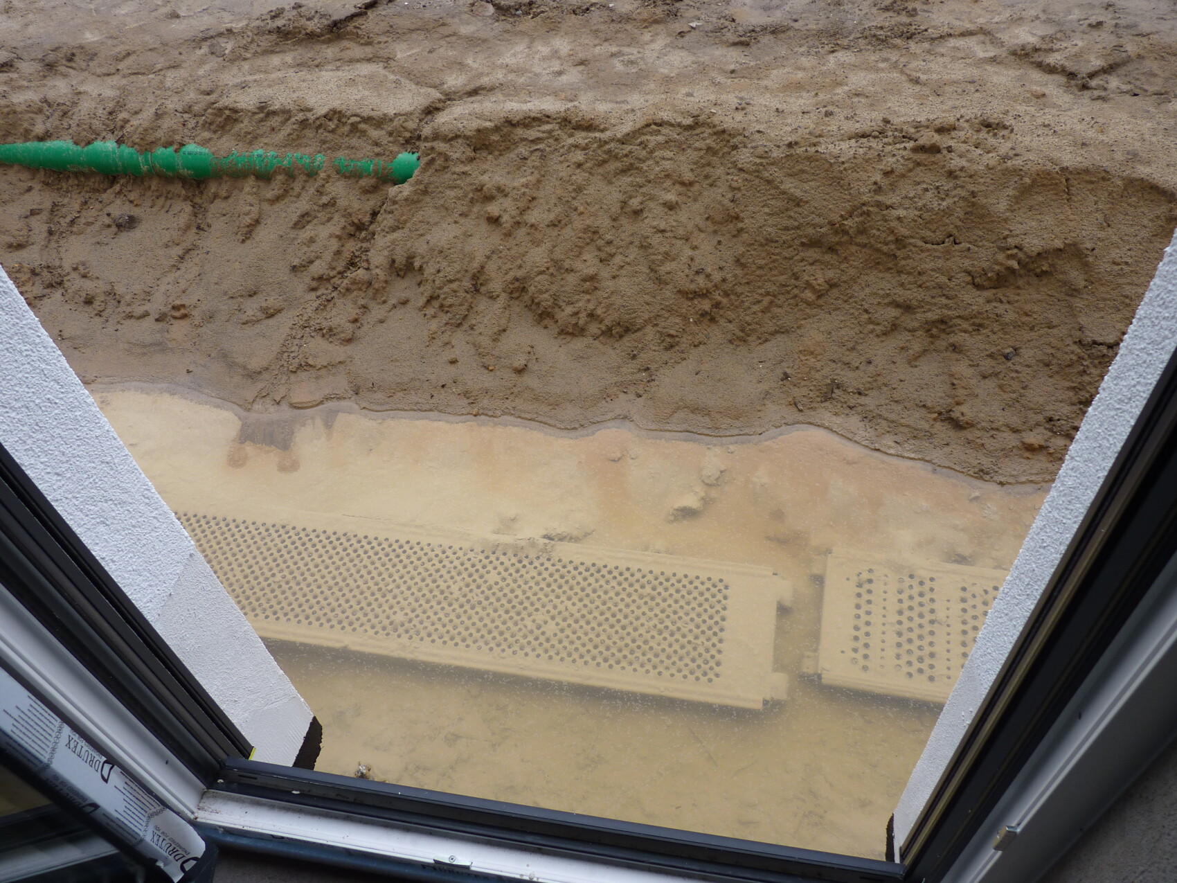 Blick durch offenes Fenster auf Baugrube, Sand und grüne Rohrleitung außerhalb des Hauses