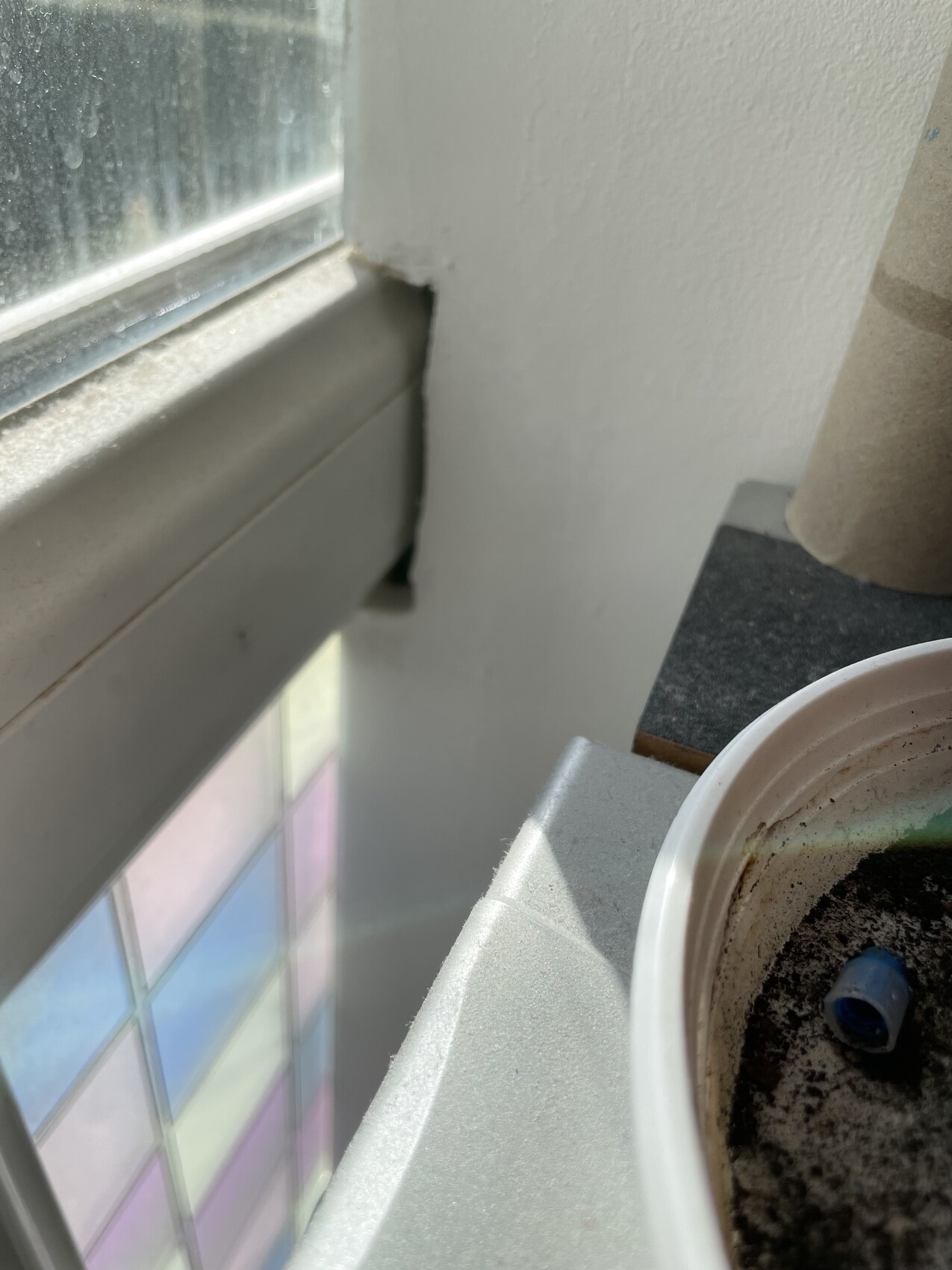 Fensterrahmen mit Spalt zur Wand; farbiges Glasfenster im Hintergrund; Blumentopf mit blauem Objekt.