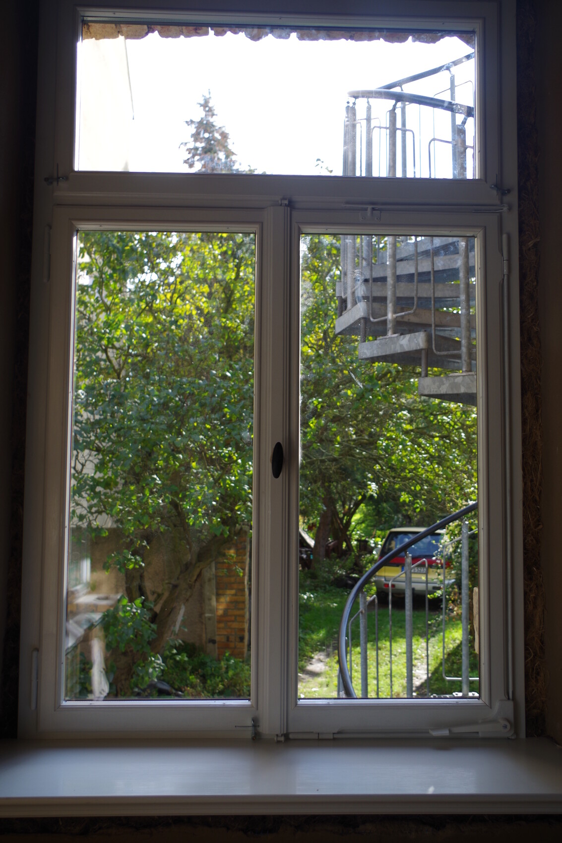 Fensterblick nach außen auf Garten mit Bäumen, rechts Metall-Außentreppe, Auto im Hintergrund