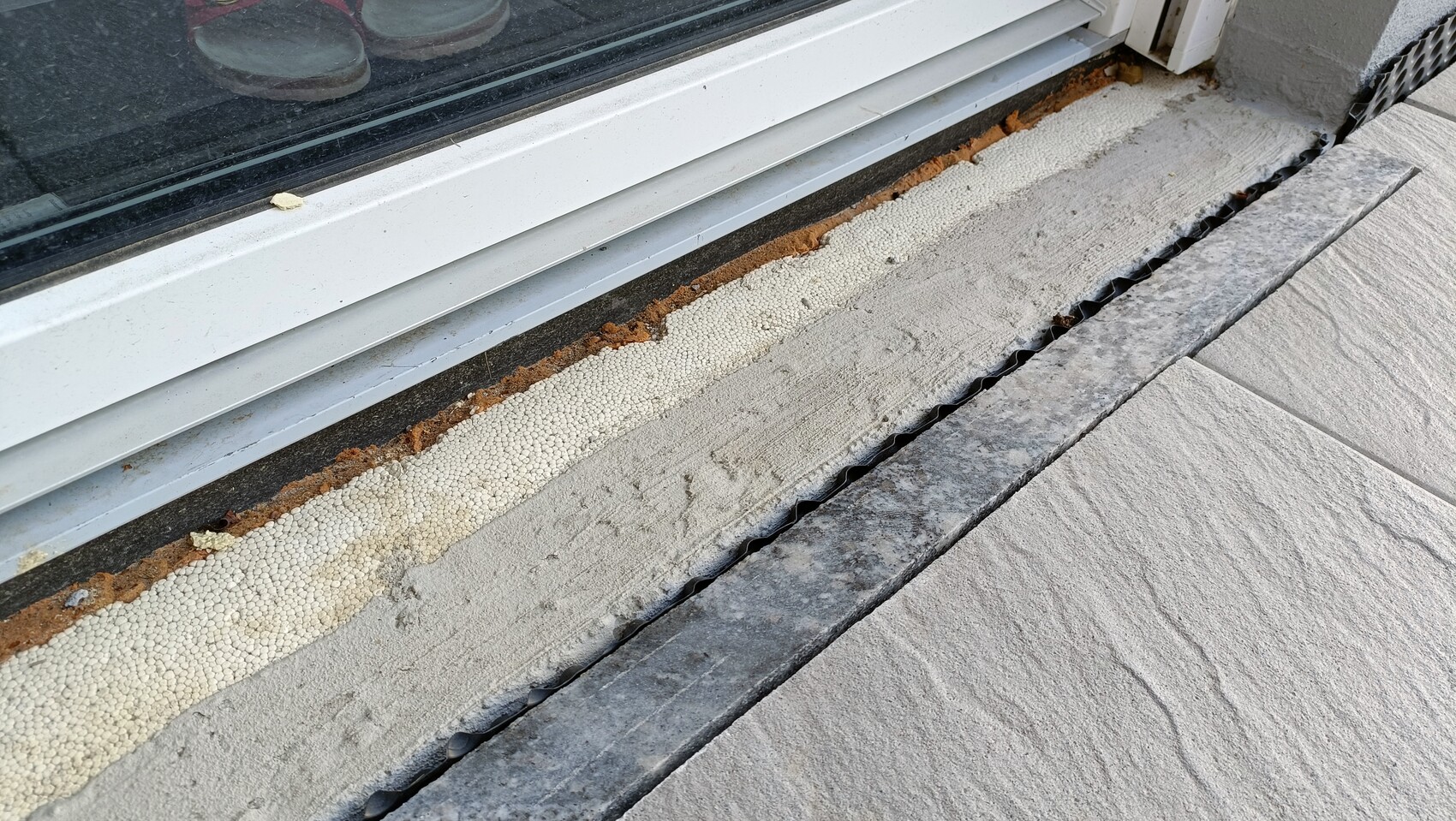 Außenansicht: Türrahmen mit Dichtung, weißem Fugen-Schaum, Rost, Steinplatten und Abflussgitter.